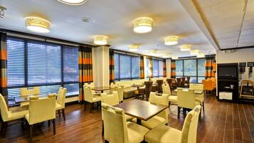 A spacious hotel dining area with multiple tables and chairs, large windows with blinds, and ceiling lights.