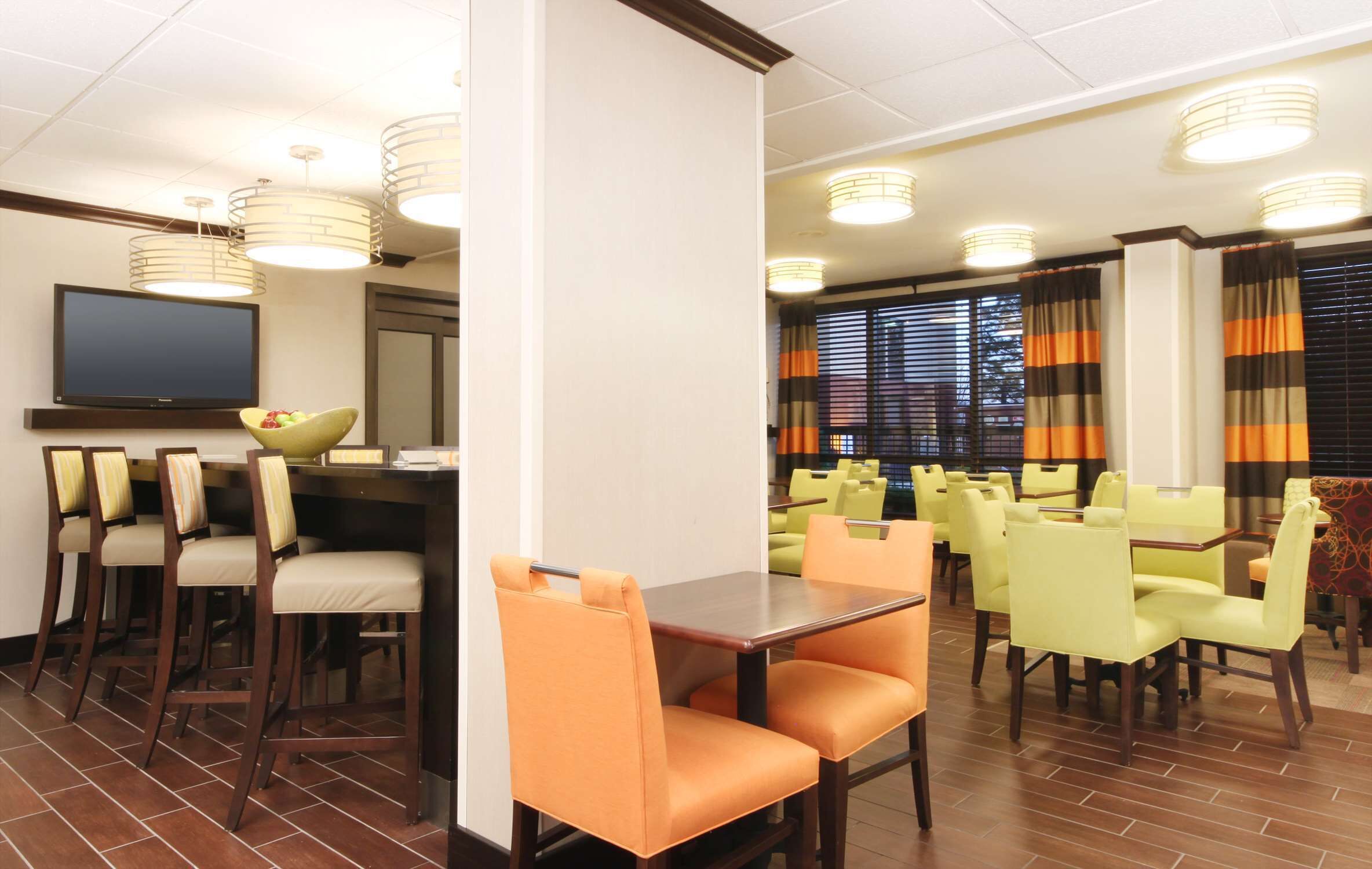 A hotel dining area with multiple tables and chairs, a flat-screen TV, and a bowl of fruit on a counter.