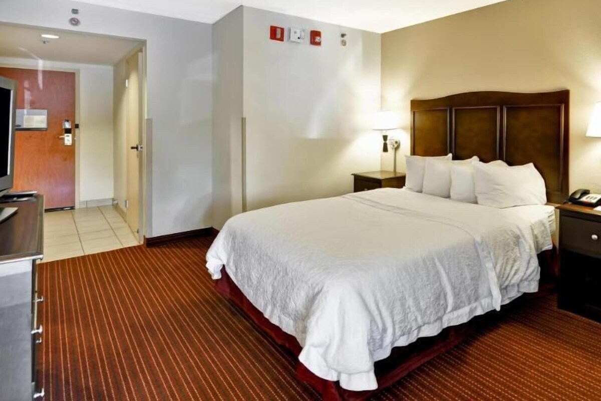 A hotel room with a large bed, a bedside table with a lamp, a desk with a chair, and a door leading to another room.