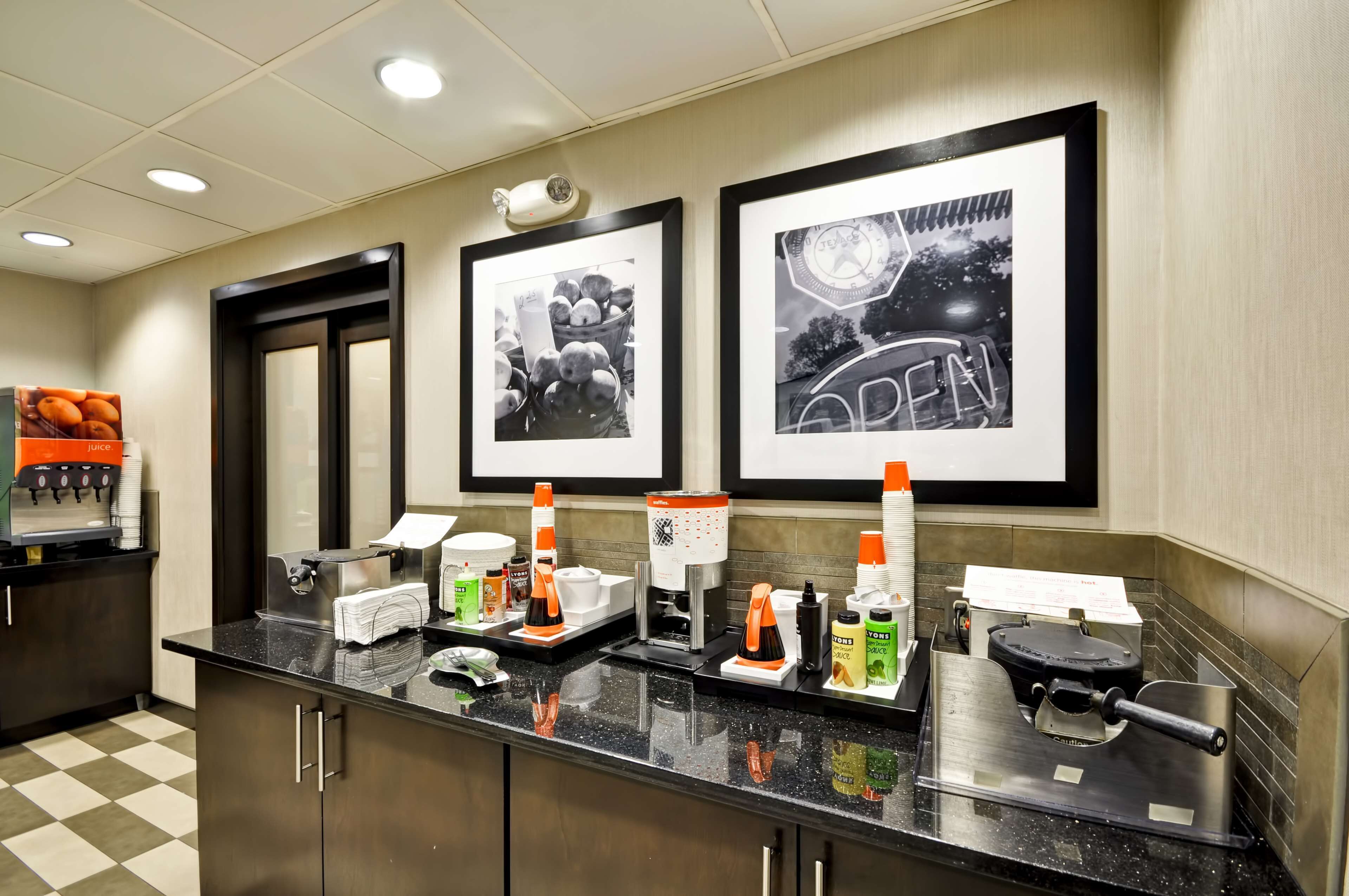 A hotel reception area with a check-in counter, framed pictures on the wall, and various amenities like a coffee machine and condiment dispensers.