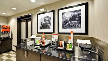 A hotel reception area with a check-in counter, framed pictures on the wall, and various amenities like a coffee machine and condiment dispensers.