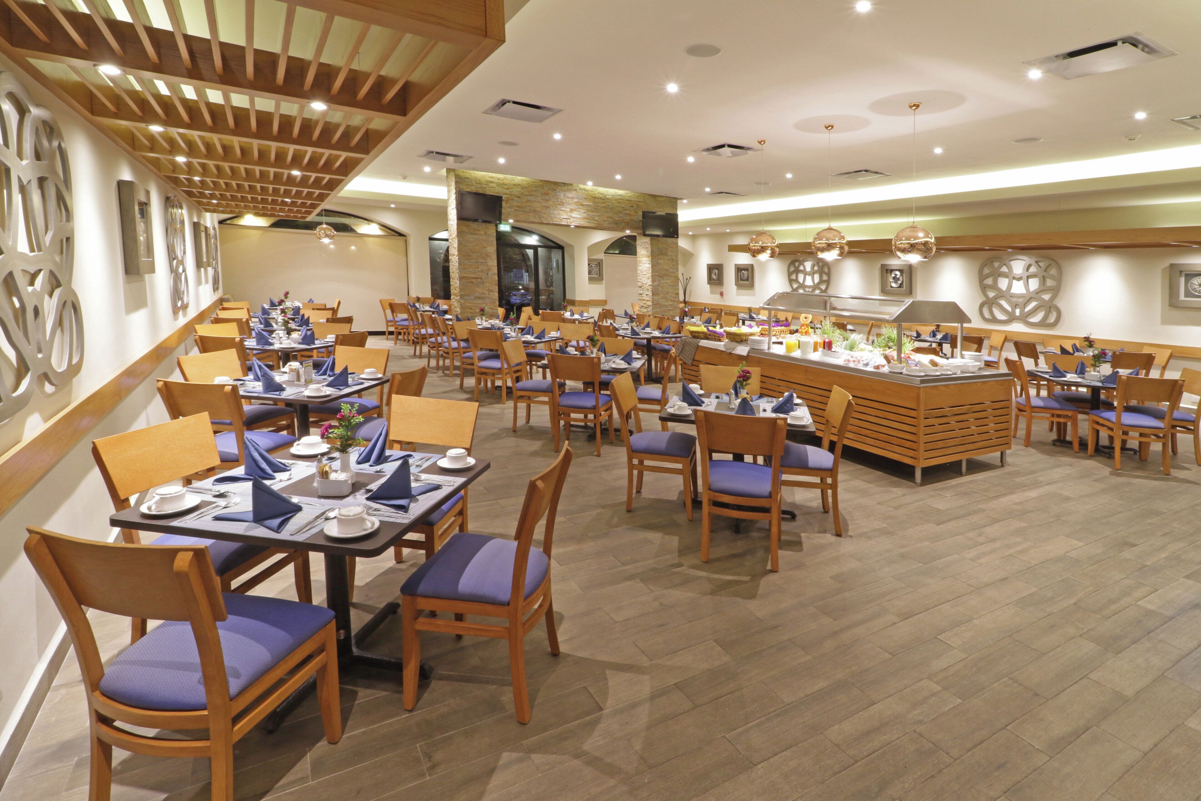 Un vaste espace dédié à la salle à manger, avec de nombreuses tables et chaises, un buffet central et un plafond haut doté d’un éclairage encastré.