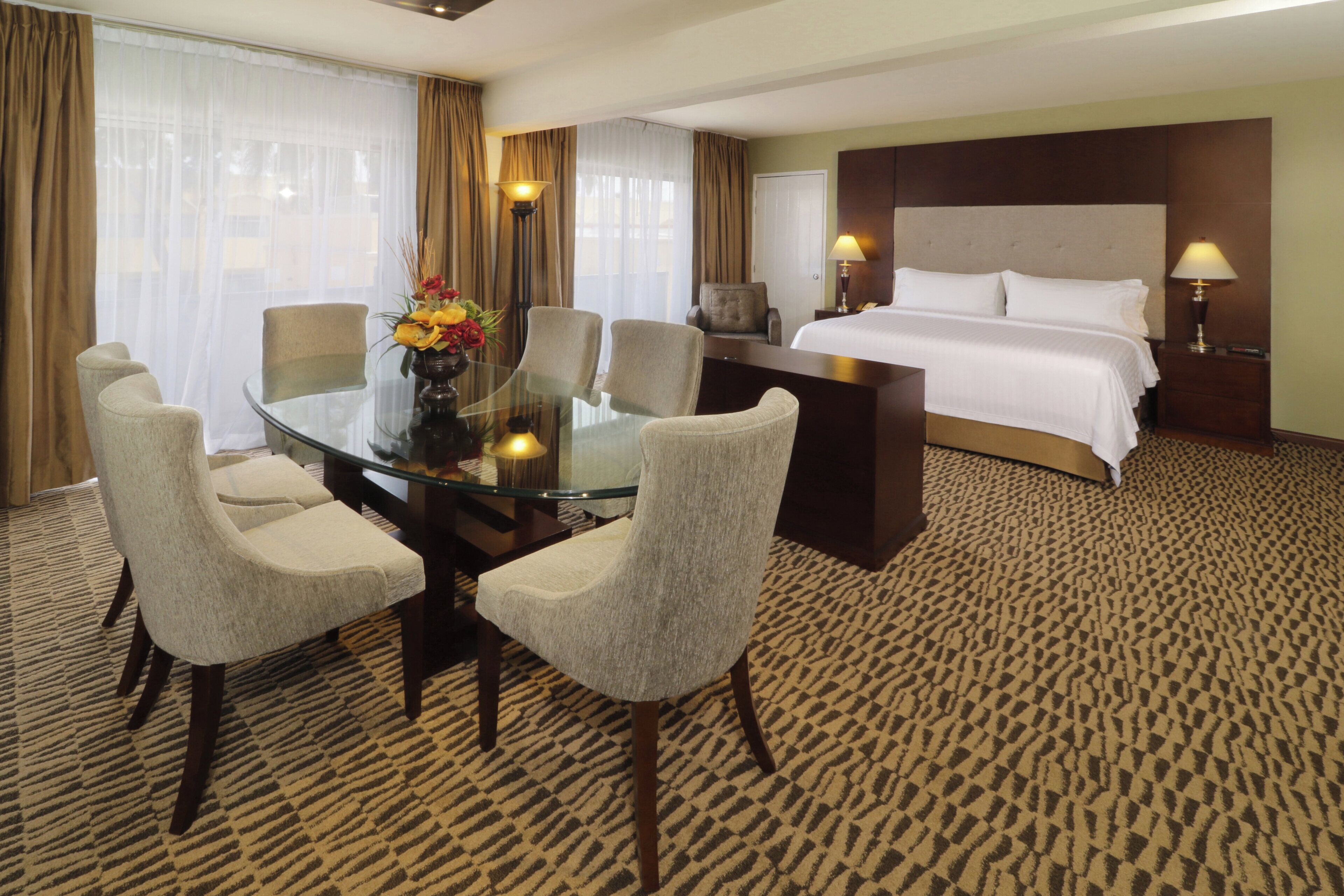 A hotel room with a glass dining table, four upholstered chairs, a bed with white linens, a nightstand with a lamp, and a large window with curtains.
