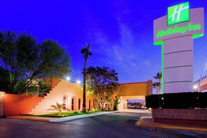 A Holiday Inn hotel entrance with a prominent sign, a parking lot, and a building with a red roof. - Holiday Inn Monterrey Norte by IHG (San Nicolas de los Garza)