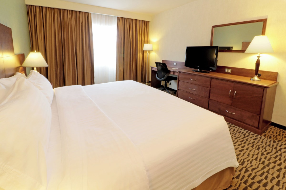 A hotel room with a large bed, a desk with a chair, a television, and two lamps.