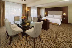 A hotel room with a glass dining table, four chairs, a bed with white linens, a headboard, two bedside lamps, and a patterned carpet.