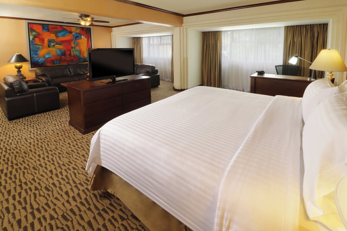 A hotel room with a large bed, a flat-screen TV, a desk with a lamp, a sofa, and a painting on the wall.