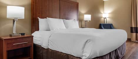 A hotel room with a large bed, wooden headboard, two bedside tables with lamps, a chair, and a nightstand with a clock.