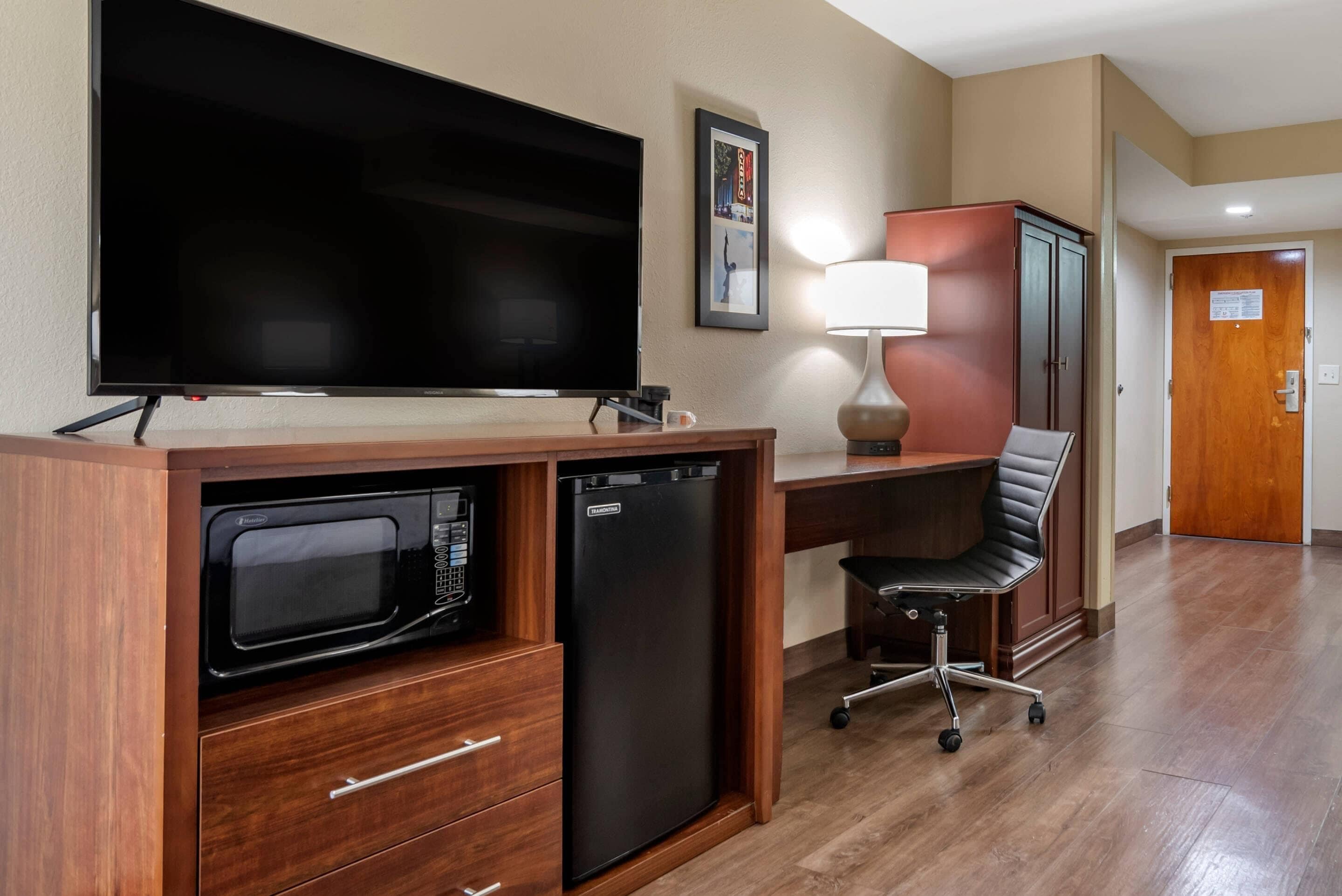 A compact hotel room with a flat-screen TV, a microwave, a mini-fridge, a desk with a lamp, a black office chair, and a wooden door.