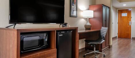 A compact hotel room with a flat-screen TV, a microwave, a mini-fridge, a desk with a lamp, a black office chair, and a wooden door.
