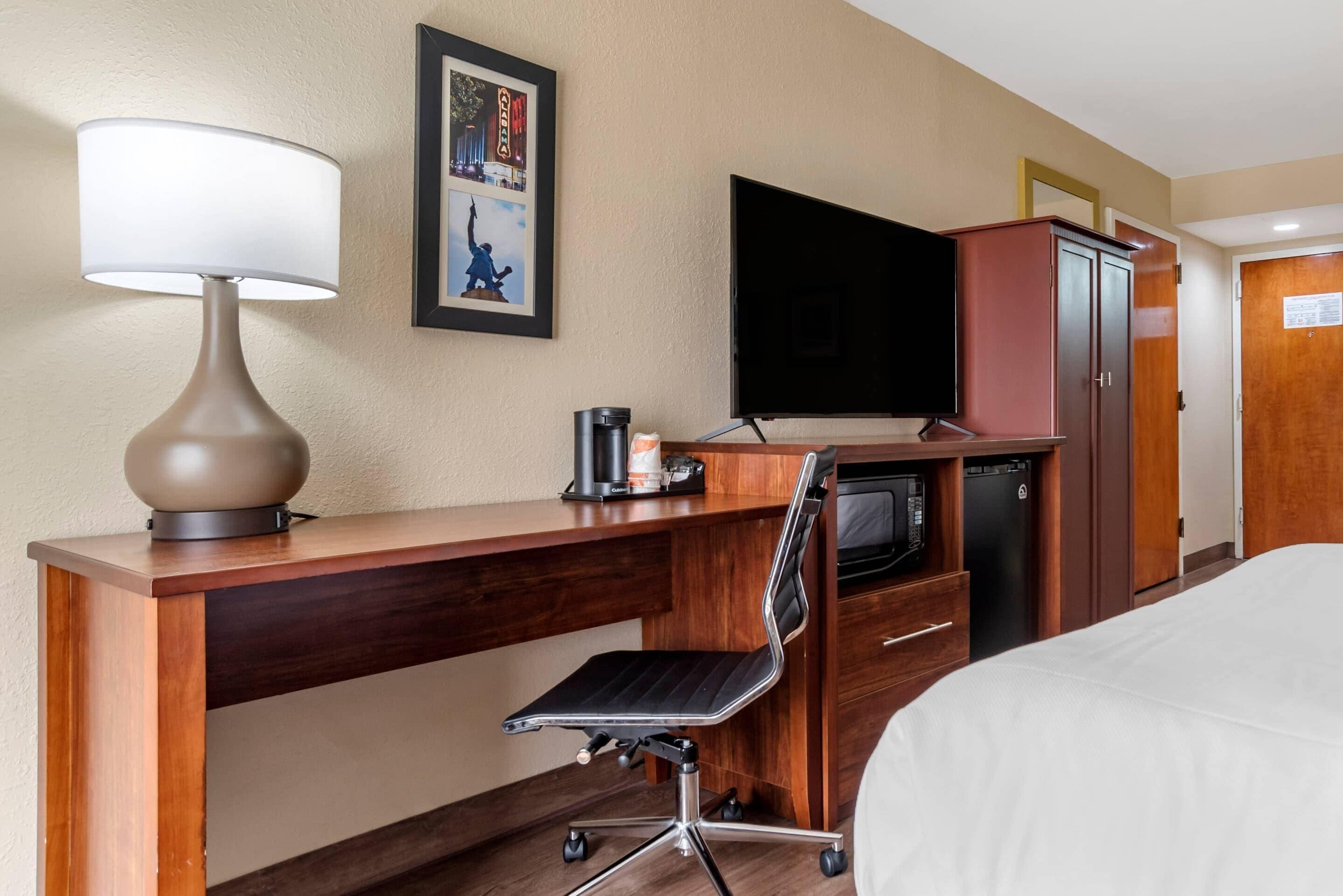 A hotel room with a wooden desk, a black office chair, a flat-screen TV, a microwave, a coffee maker, and a bed with white bedding.