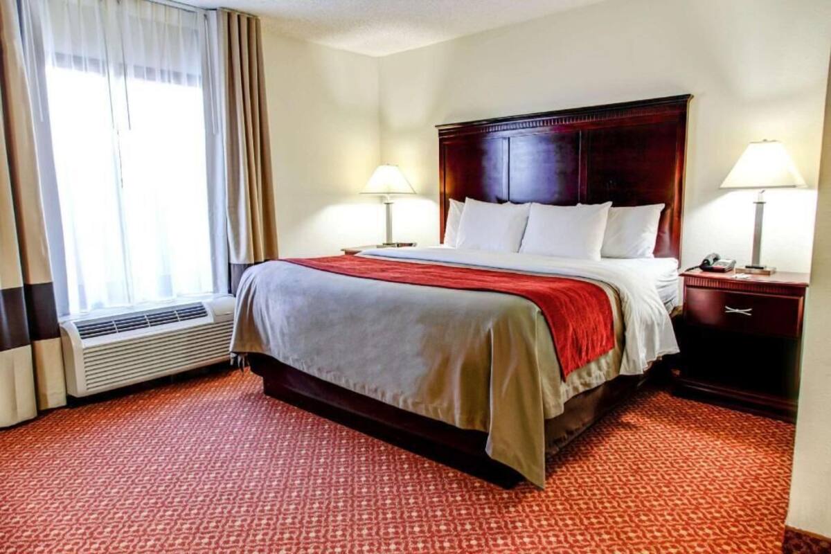 Ein Hotelzimmer mit einem großen Bett, zwei Nachttischen mit Lampen, einem Fenster mit Vorh ängen und einem Teppichboden.