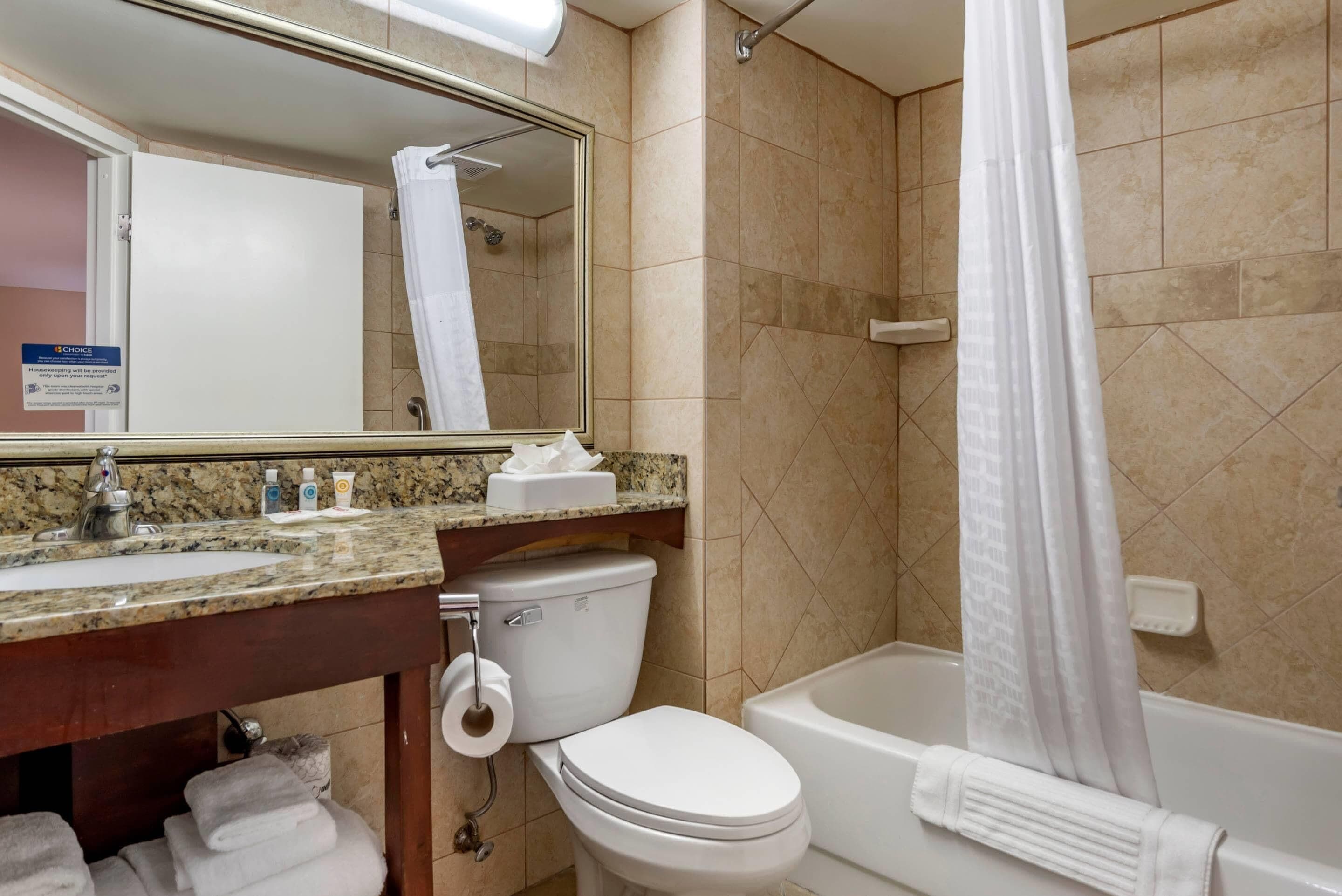 Un baño con lavamanos, inodoro y bañera. Hay un perchero con toallas, un espejo y un cartel en la pared.