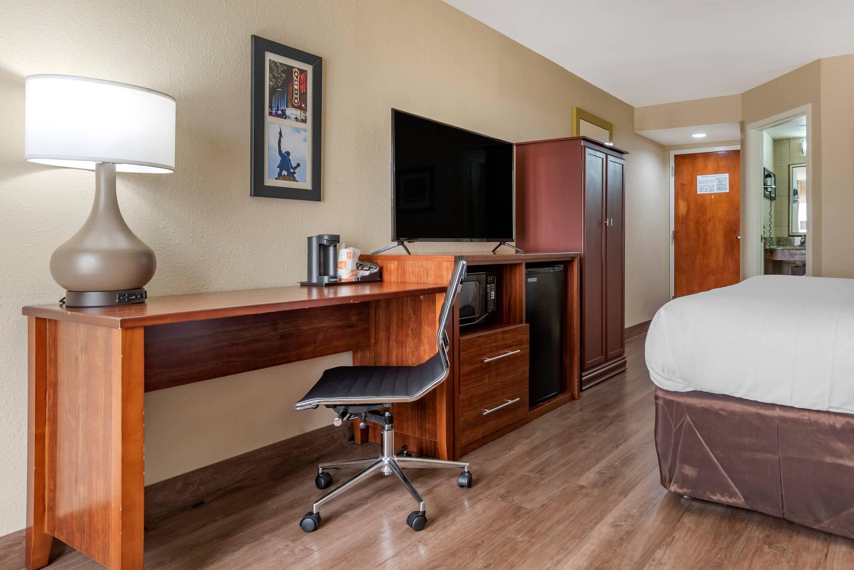 A hotel room with a wooden desk, a black office chair, a television, a minibar, a wardrobe, and a bed.