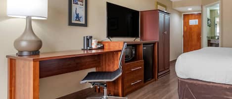 A hotel room with a wooden desk, a black office chair, a television, a minibar, a wardrobe, and a bed.