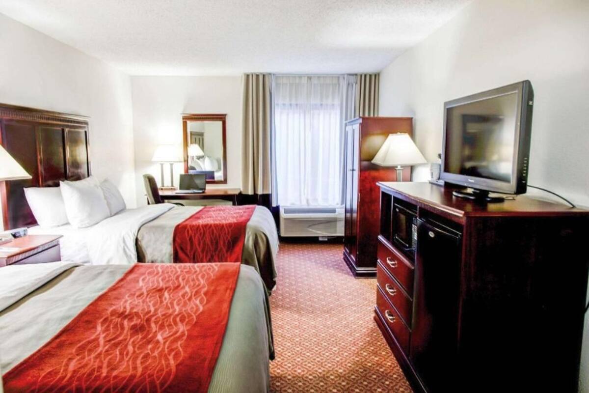 A hotel room with two beds, a TV, a desk with a lamp, a mirror, and a window with curtains.