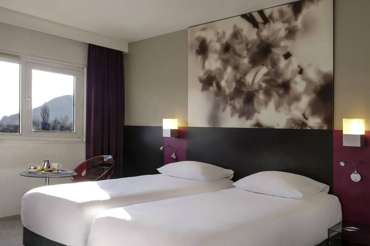 Une chambre d’hôtel avec deux lits, une grande peinture abstraite au mur, une petite table avec des éléments pour le petit-déjeuner et une vue sur l’extérieur par la fenêtre.