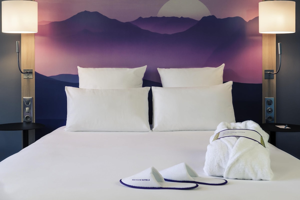 A hotel room with a bed, pillows, a nightstand, a lamp, a robe, slippers, and a towel.