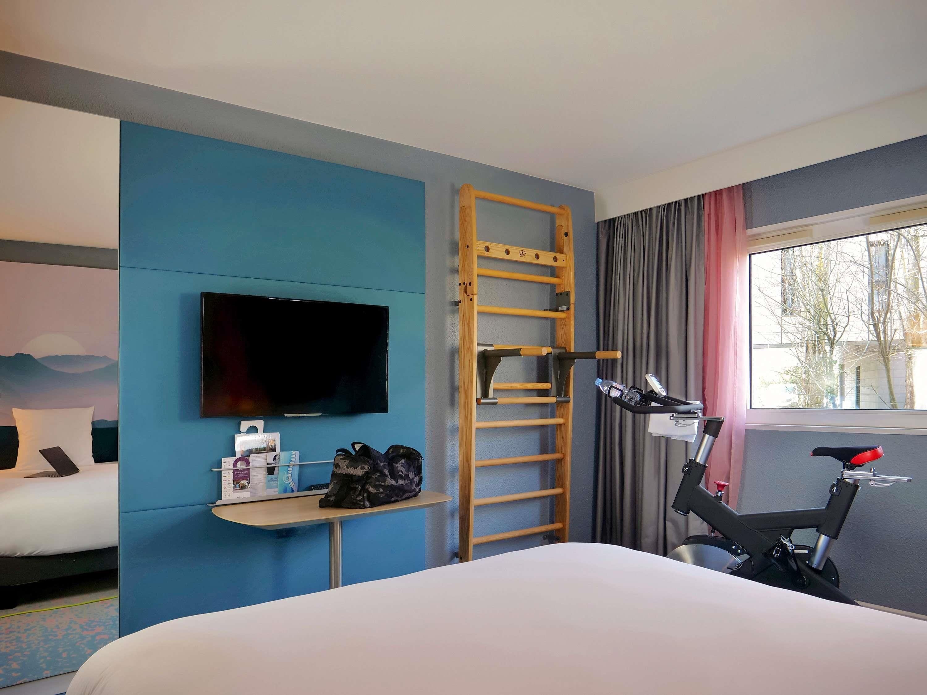 A hotel room with a bed, a TV, a desk with a laptop, a bicycle, and a window with curtains.