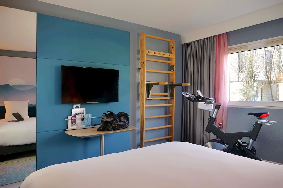 A hotel room with a bed, a TV, a desk with a laptop, a bicycle, and a window with curtains.