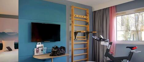 Habitación de hotel con cama, televisor, escritorio con una computadora portátil, bicicleta y ventana con cortinas.