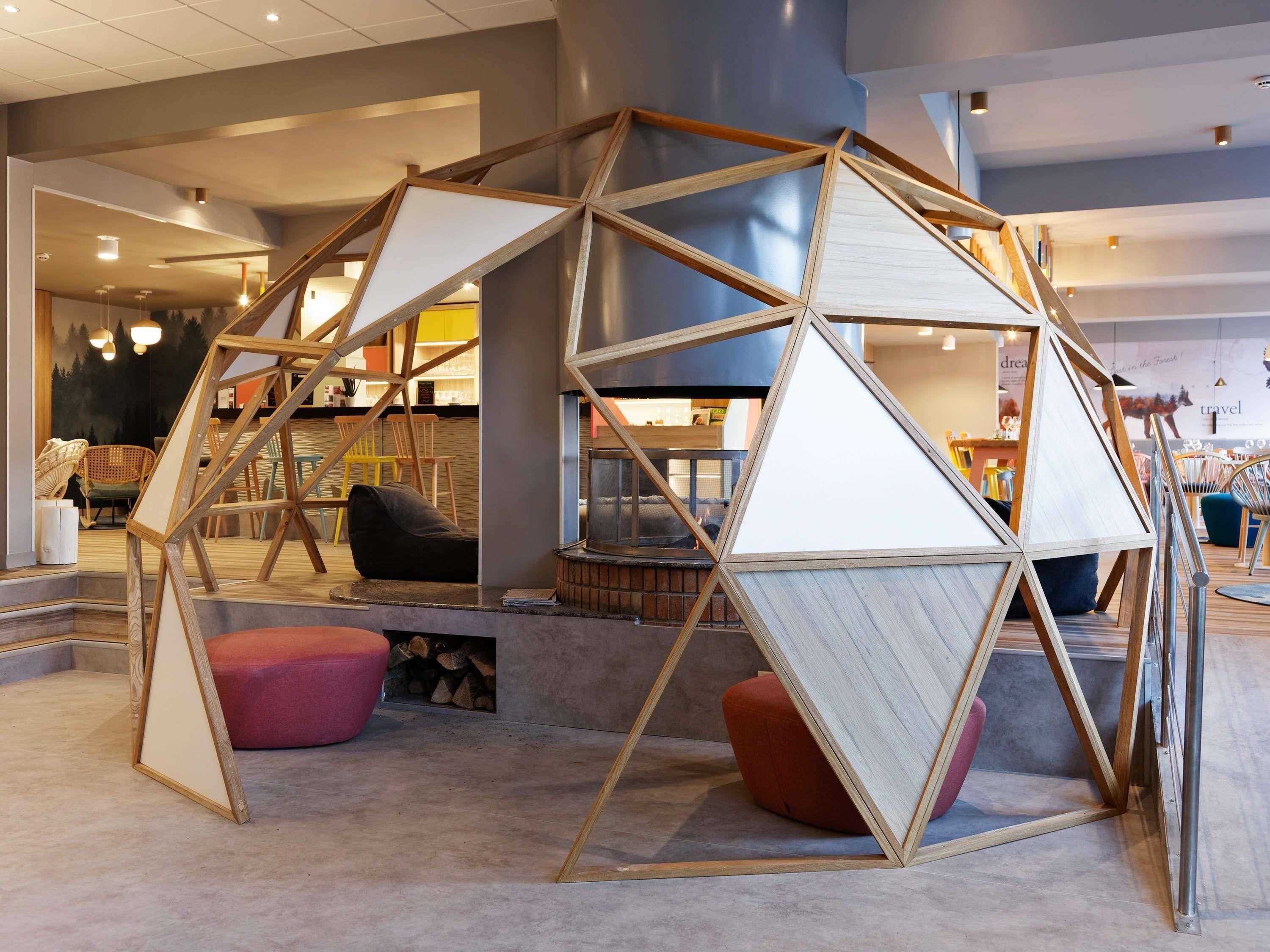 Ein modernes Interieur mit einer geometrischen Holzkonstruktion, Sitzbereichen und einem sichtbaren Kamin.