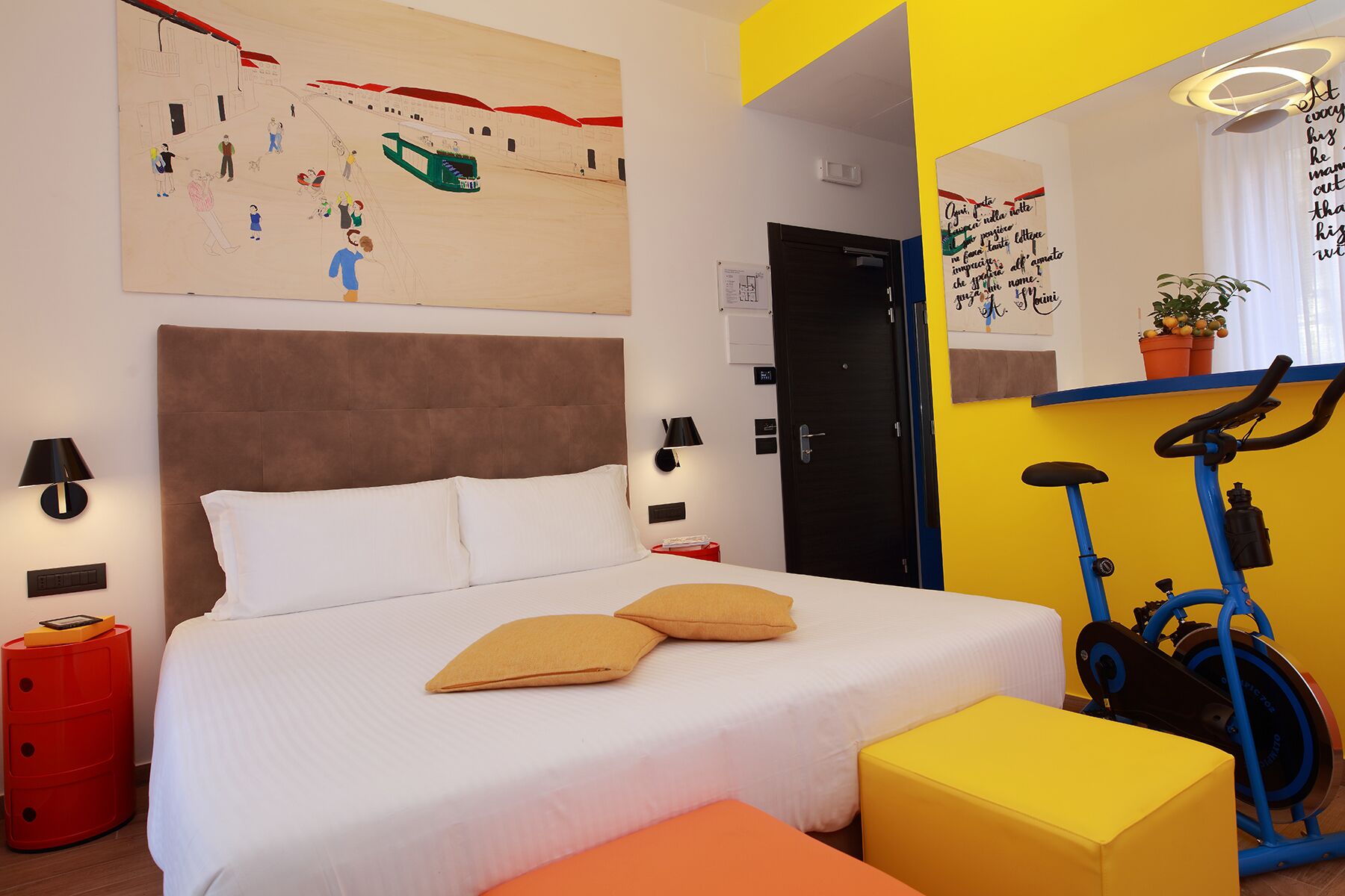 A bedroom with a bed, a red nightstand, a yellow ottoman, a blue exercise bike, and a large mirror.