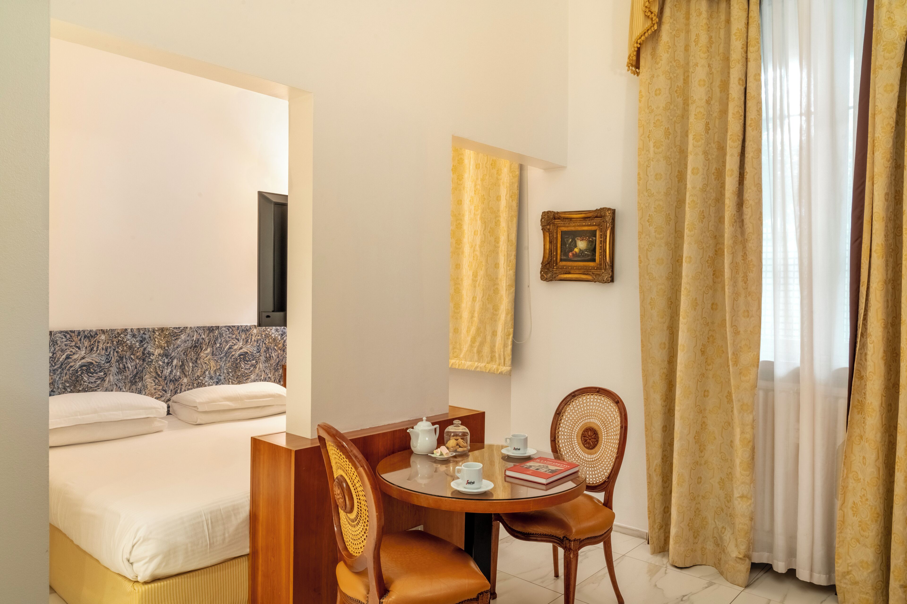 Una camera d'albergo con un letto, un tavolino con teiera, tazze e un libro, una sedia e un quadro appeso al muro.