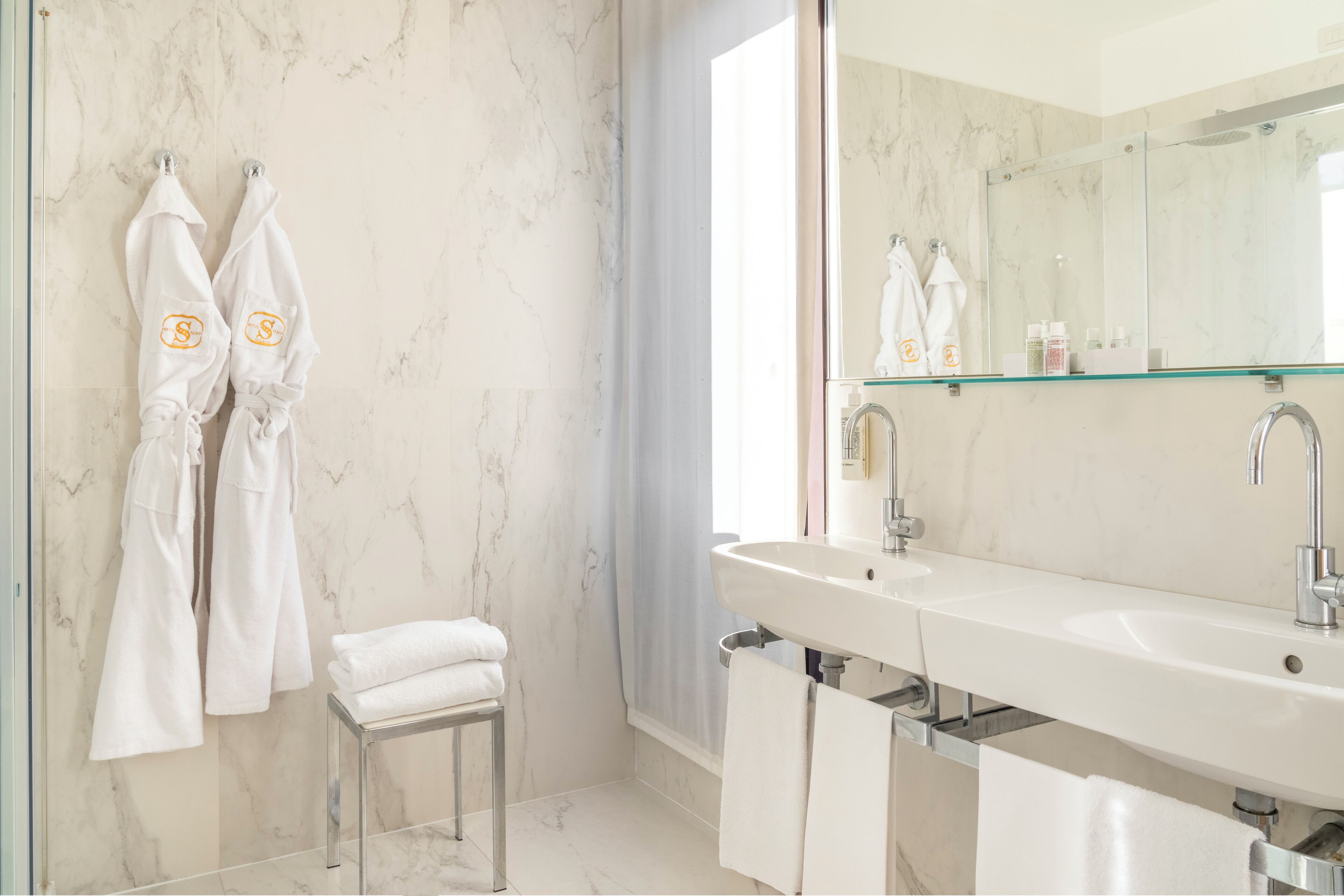 A modern bathroom with a marble wall, a large mirror, a sink, and two white towels on a stool.