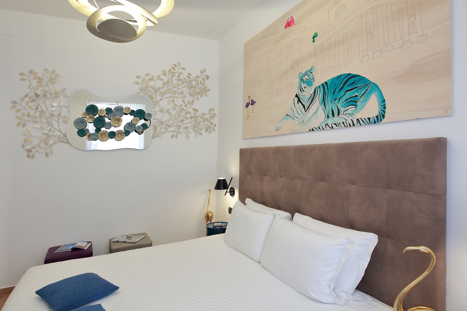 Une chambre d’hôtel avec un grand lit, un miroir orné de motifs végétaux, une peinture murale représentant un tigre et un luminaire moderne.