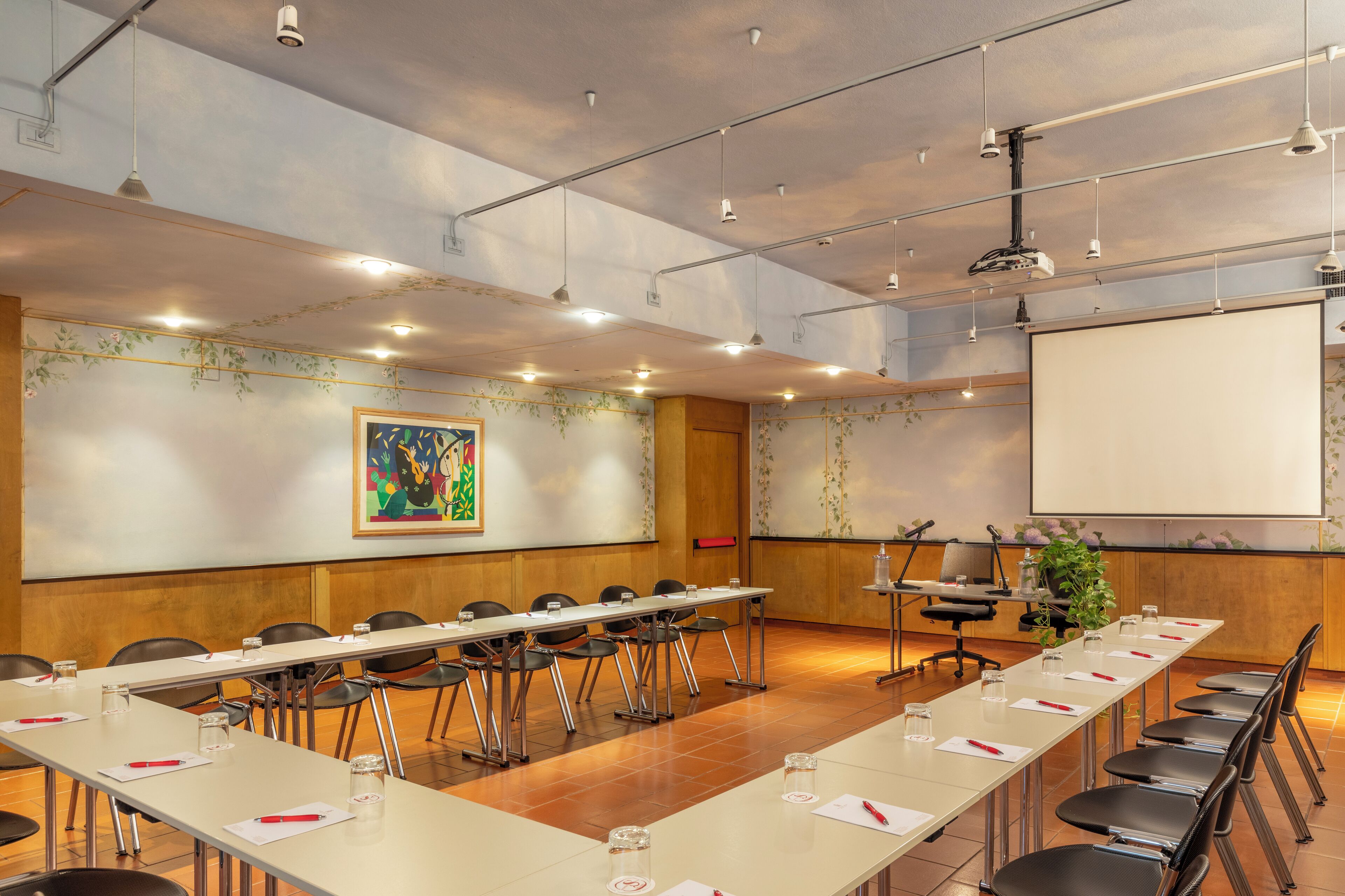A conference room with a long table, chairs, a projector screen, and a large painting on the wall.
