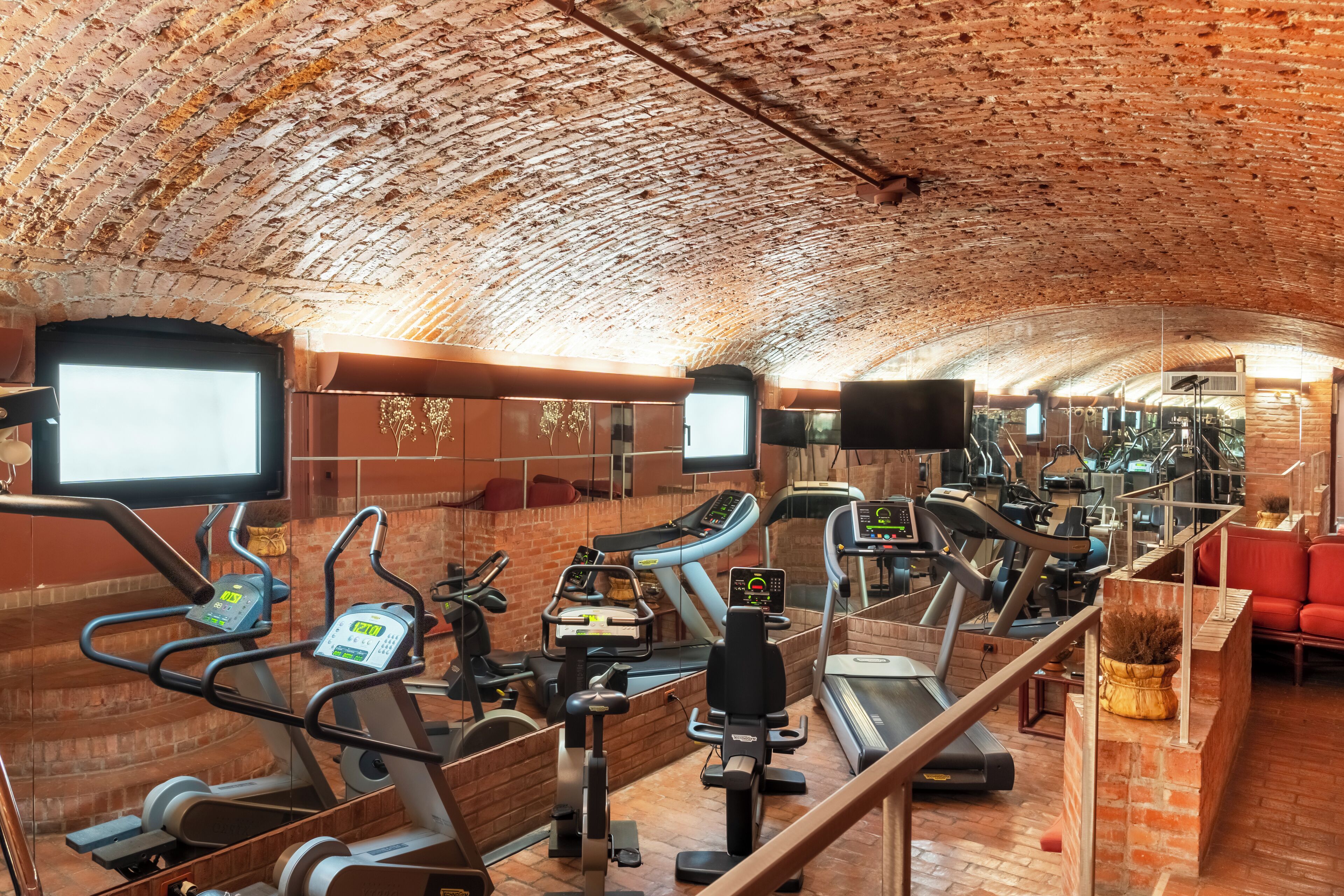A gym with various cardio machines, a TV, and a brick ceiling.