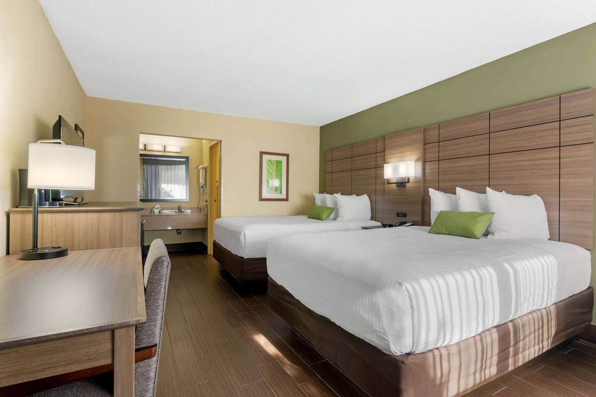Habitación de hotel con dos camas, un escritorio, una silla, un espejo y un baño visible a través de una puerta abierta.