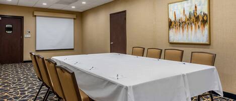 Un salón de conferencias con una larga mesa, sillas, una pantalla de proyector y un cuadro en la pared.