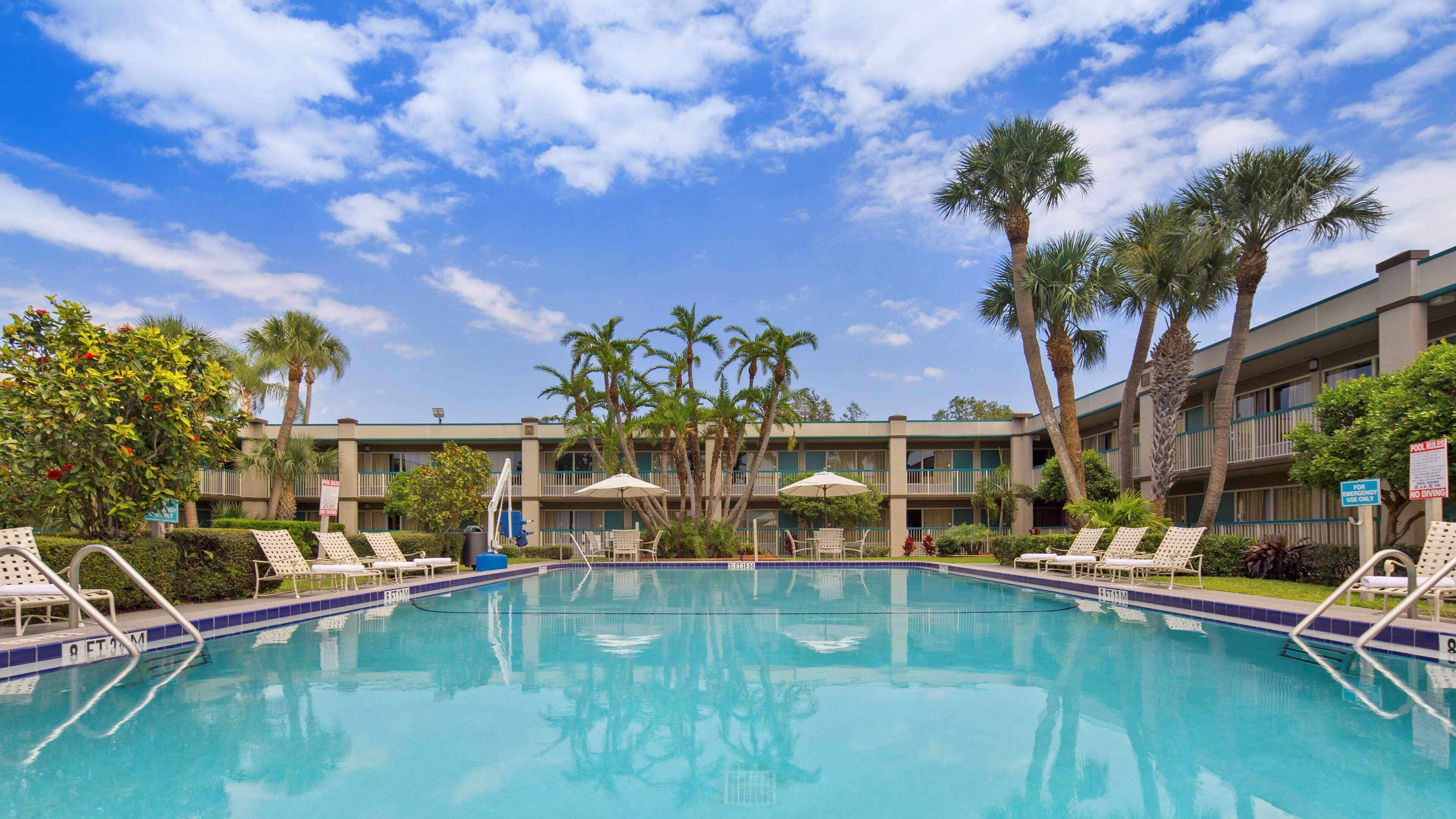 A swimming pool with lounge chairs and palm trees, surrounded by a multi-story building with balconies.