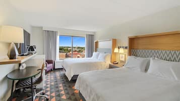 Quarto de hotel com duas camas, uma escrivaninha com cadeira, janela com vista e uma mesinha de cabeceira com abajur.