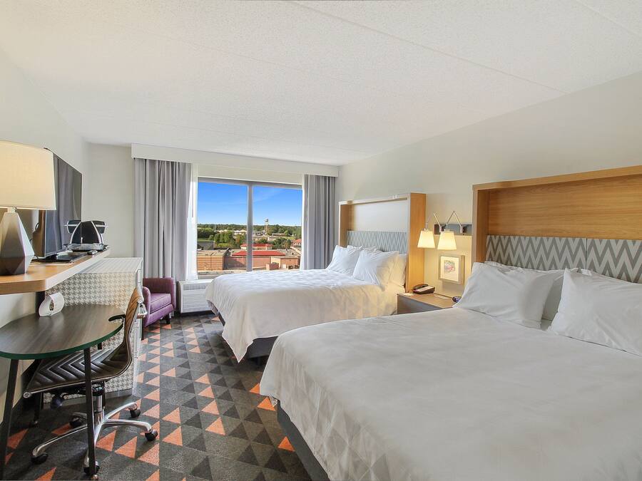 Quarto de hotel com duas camas, uma escrivaninha com cadeira, janela com vista e uma mesinha de cabeceira com abajur.