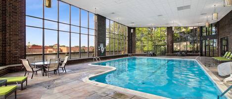An indoor swimming pool with clear glass walls, stone flooring, and outdoor seating areas.