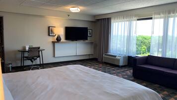 A hotel room with a bed, a TV, a sofa, a desk with a chair, and a window with curtains.