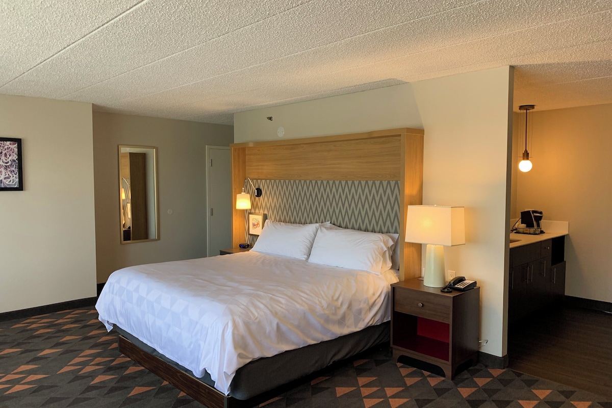 A hotel room with a bed, bedside table, lamp, and a small desk with a coffee machine.