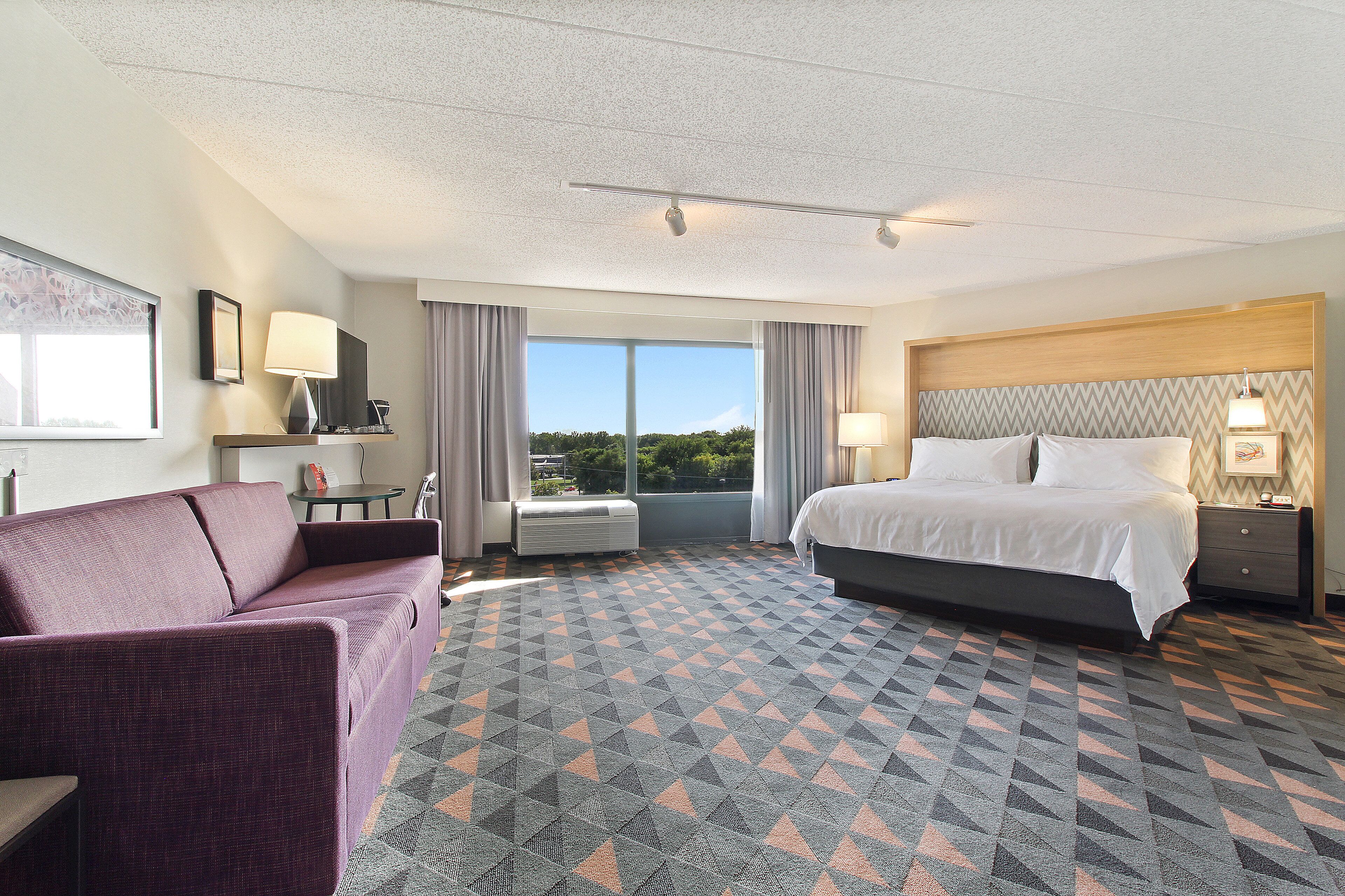 A hotel room with a bed, a chair, a desk with a lamp, and a large window offering a view.