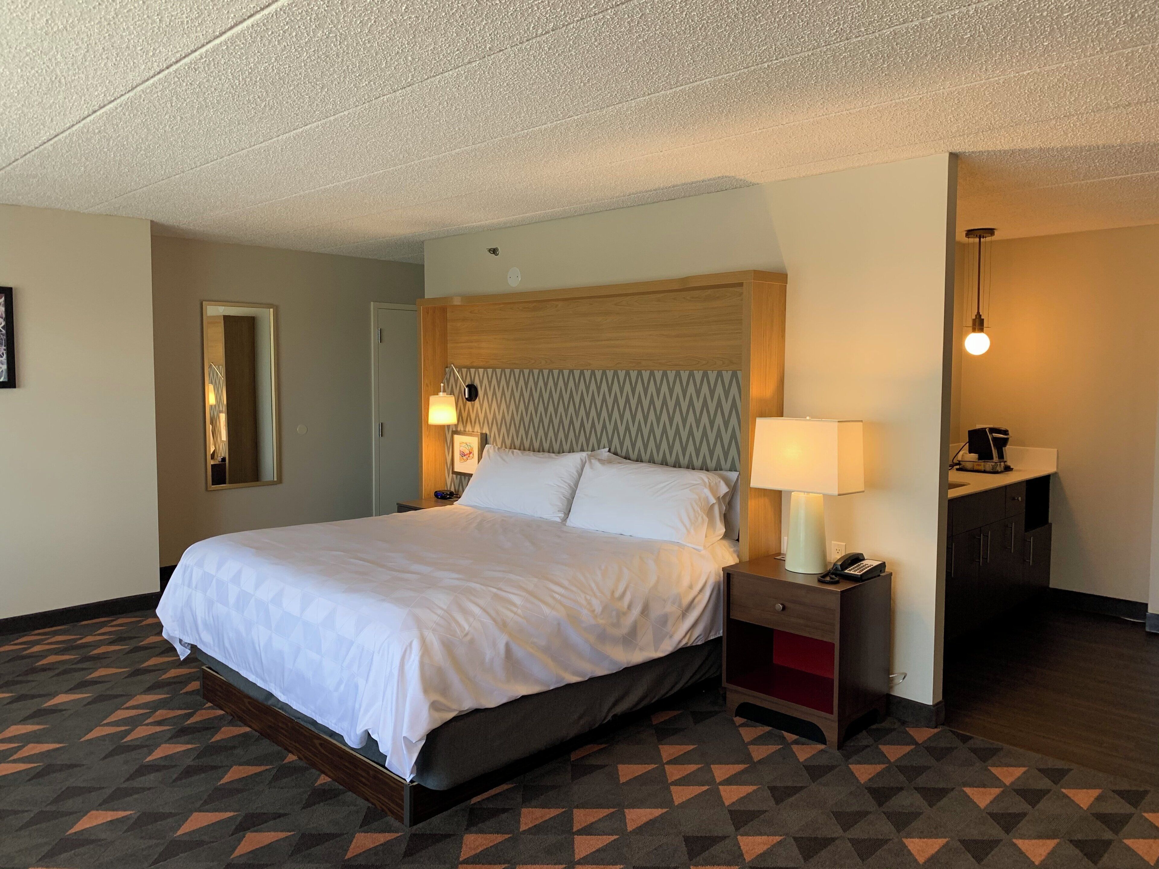 A hotel room with a bed, bedside lamps, a nightstand, a mirror, and a wall-mounted light fixture.