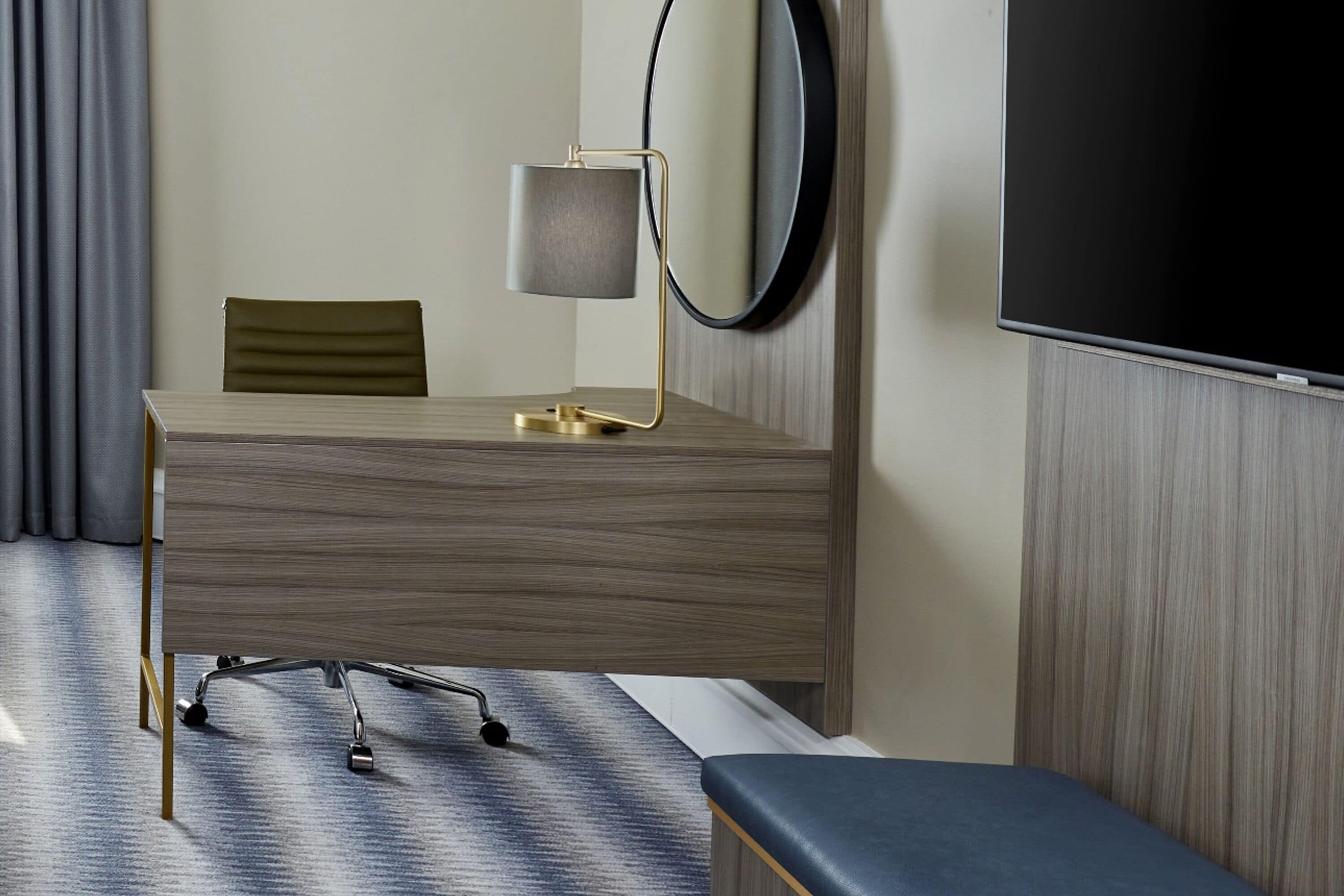 A hotel room with a wooden desk, a chair, a bedside table, a TV, and a large mirror.