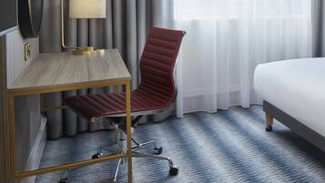 Een hotelkamer met een bed, een bureau met een lamp, een stoel en uitzicht op het landschap door dunne gordijnen.