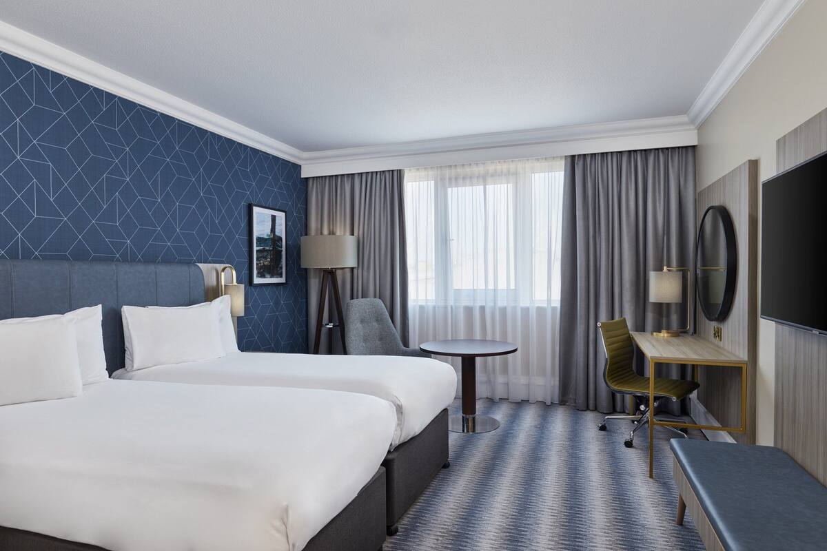 Une chambre d’hôtel avec deux lits, un bureau, une chaise, une télévision et une fenêtre avec des rideaux.