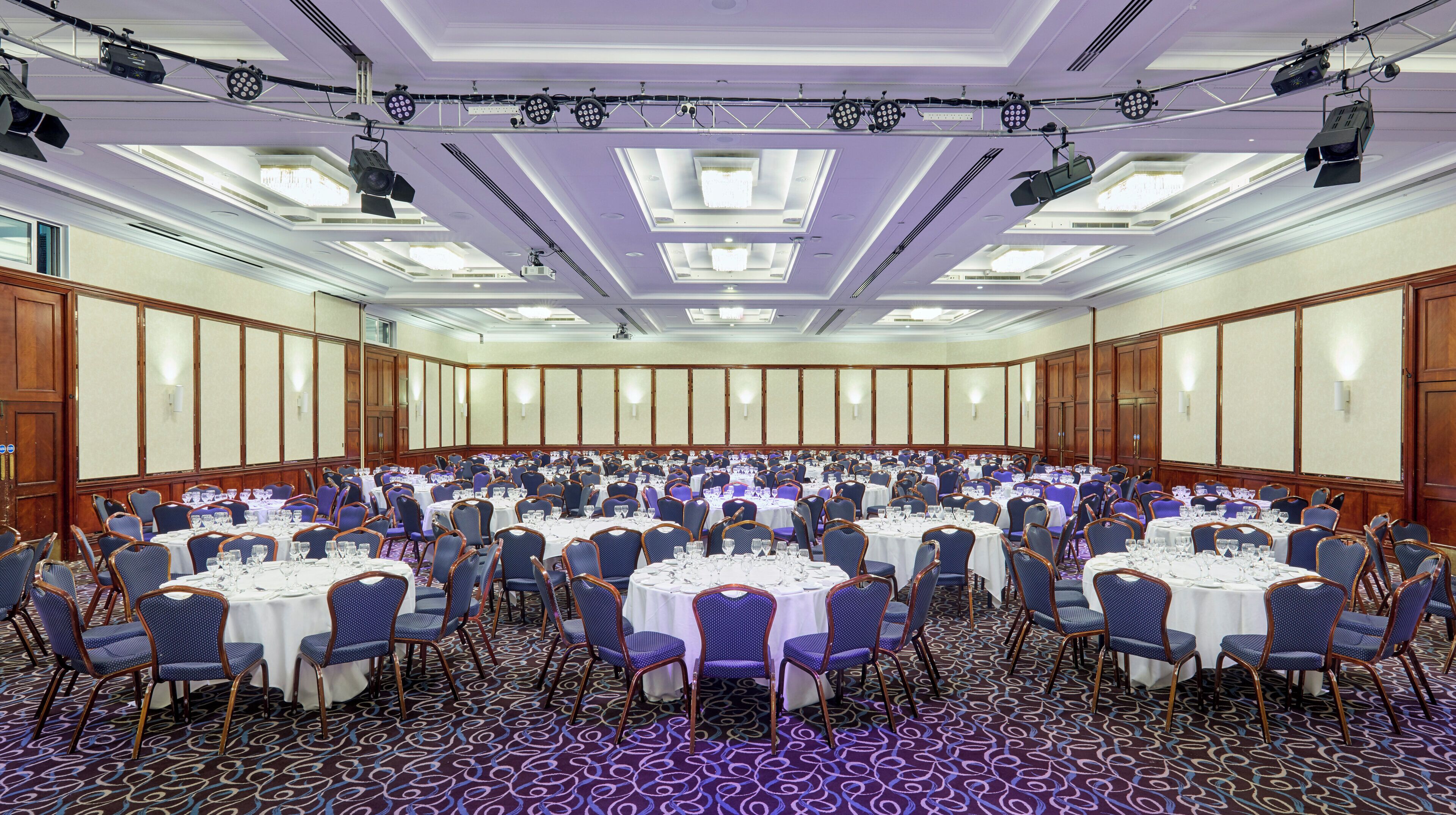 Une grande salle de conférence dotée de tables rondes et de chaises, et équipée de projecteurs et d’un système d’éclairage installés au plafond.