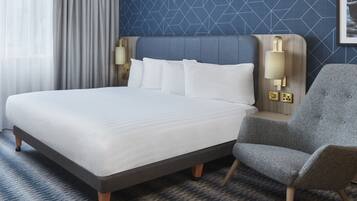 A hotel room with a large bed, a grey armchair, a headboard with wall-mounted lamps, and a patterned blue wall.