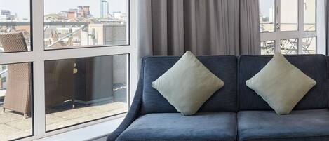 Una habitación de hotel moderna con un ventanal grande, un sofá azul oscuro con dos cojines y una mesa redonda de madera.
