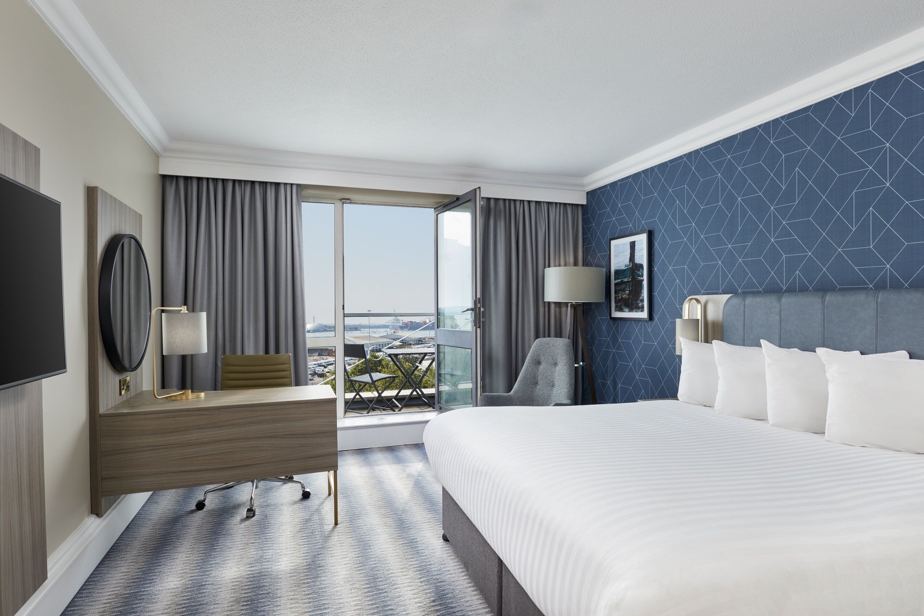 A hotel room with a large bed, a desk with a chair, a flat-screen TV, and a large window with a view of the city.