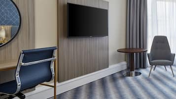 Una habitación de hotel con un televisor de pantalla plana montado en la pared, una silla azul, una mesa redonda y una alfombra estampada.
